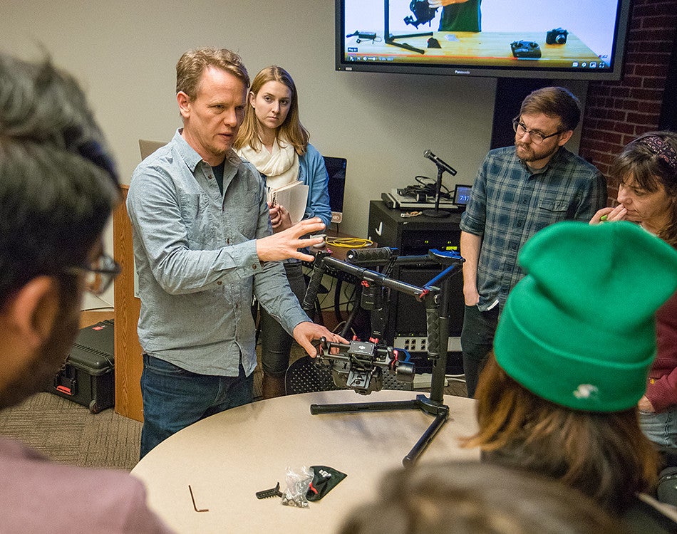 Wes Pope demonstrates how to use camera equipment to a group of students surrounding him