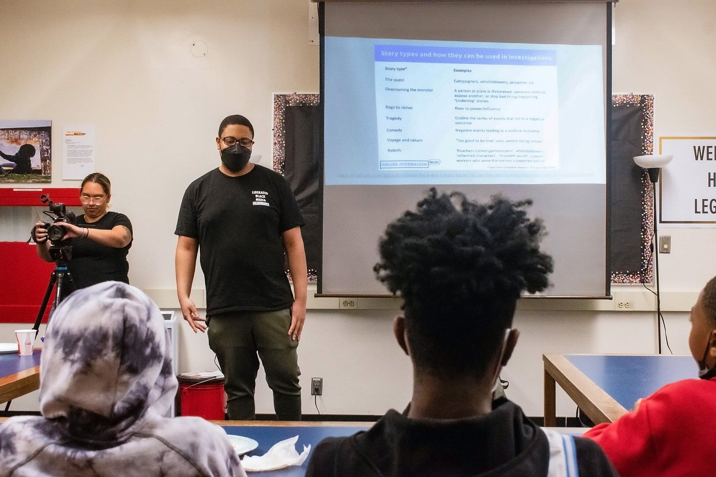 Bruce Poinsette stands in front of a classroom of students with a projector screen behind him