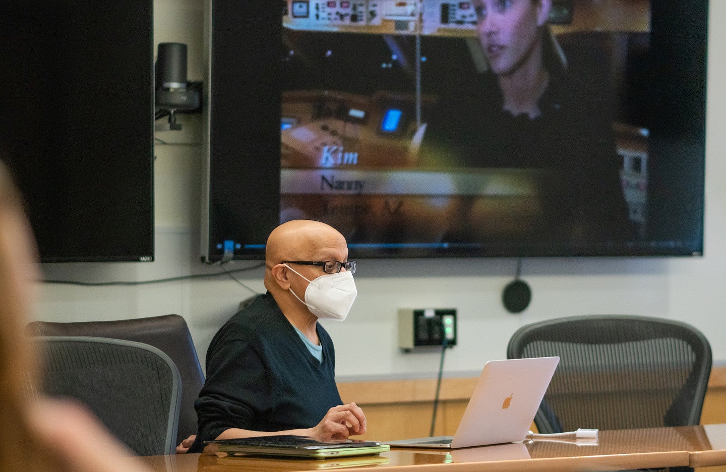 Bish Sen wears a mask and uses a laptop in front of a large tv screen