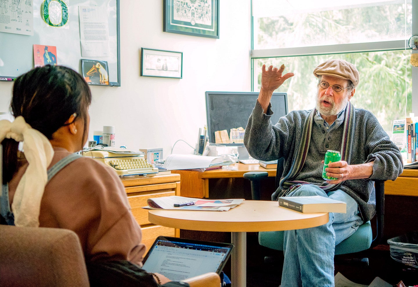 Peter Laufer sits in a desk chair with his back to windows and gestures to a student who sits across a table from him