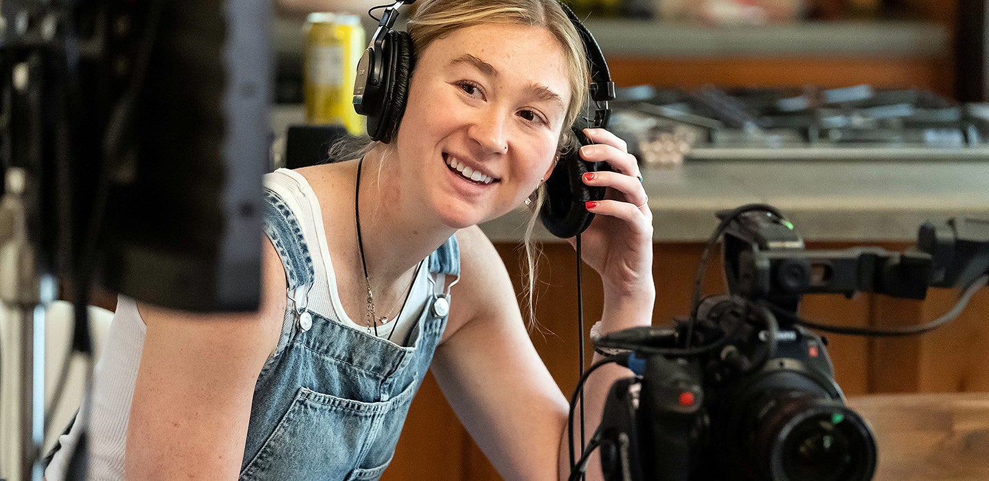 a student using camera equipment adjusts headphones while smiling at a monitor 