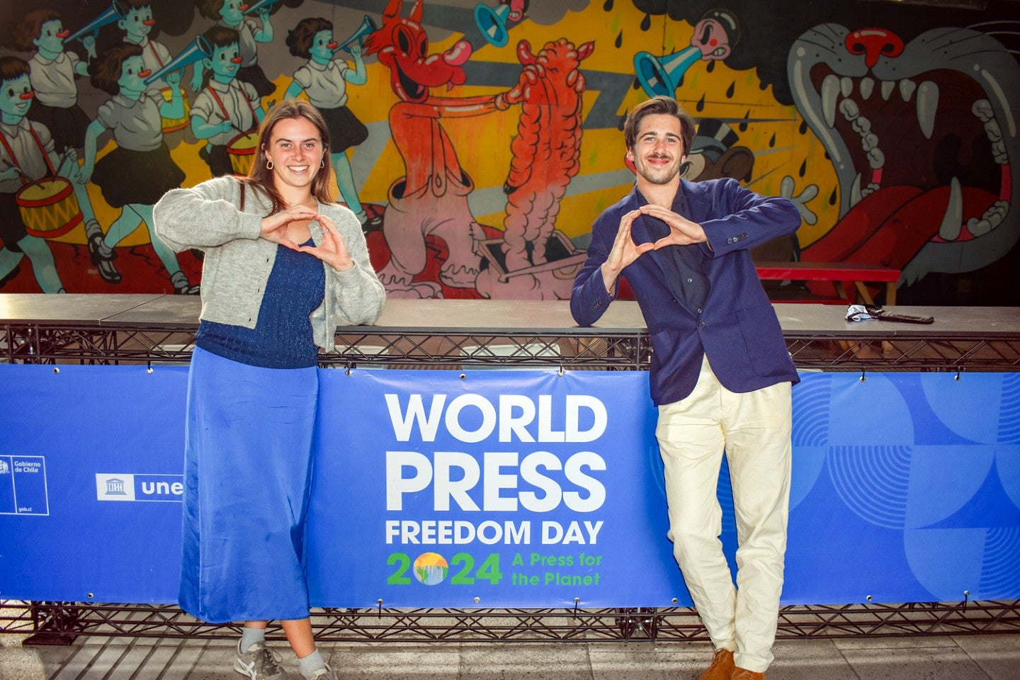 Ruby Wool and Zachary Jones Neuray, student journalists representing the SOJC at the World Press Freedom Day conference, make O shapes with their hands while standing in front of a bright blue banner and a colorful mural