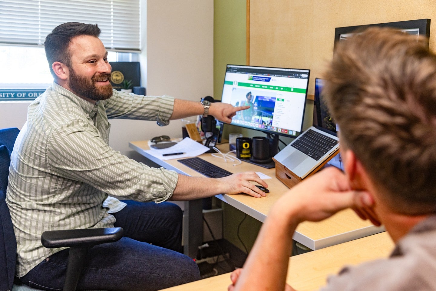 Henry Wear sits at his computer and gestures to his computer screen while talking to a student about sports PR jobs