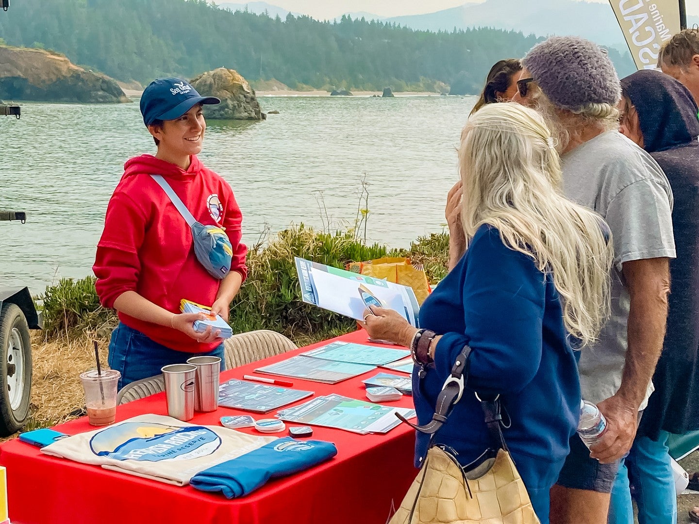 Sof Fox, a UO science communication minor, stands at a table laid out with informational materials with a river and pine forest in the background