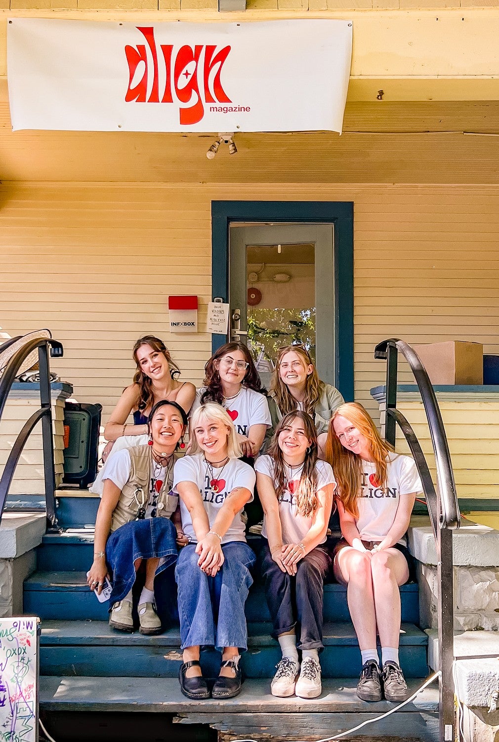 seven student members of the Align Magazine staff sit on the front stairs of a house underneath a banner for Align