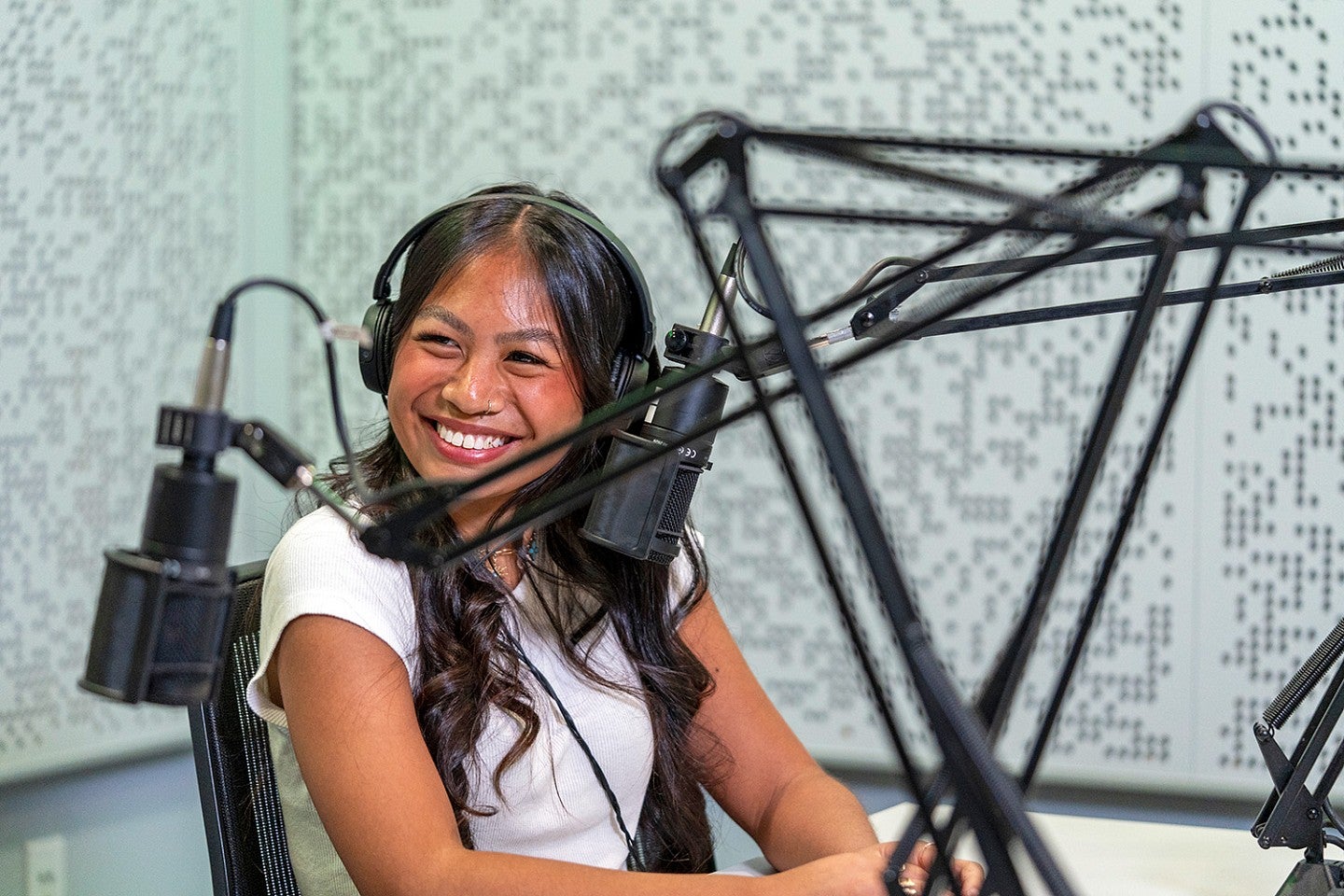 a student looks off camera and smiles while sitting in a podcast recording studio and wearing headphones