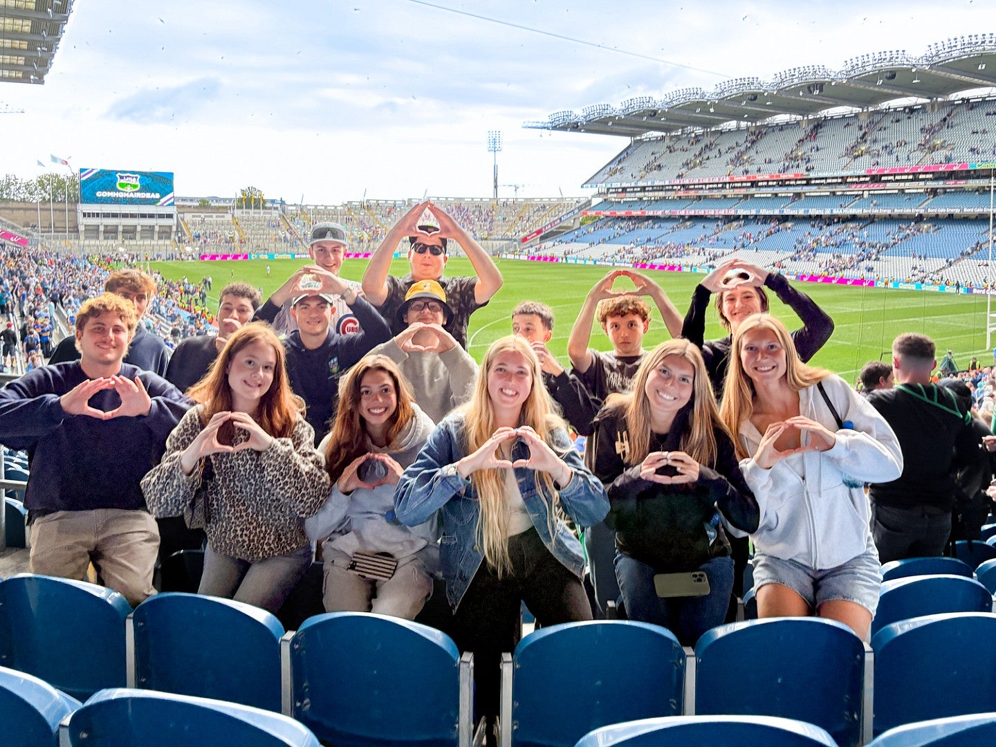a group of University of Oregon School of Journalism and Communication students throw their Os as they pose in the stands of a large athletic stadium, with the field behind them