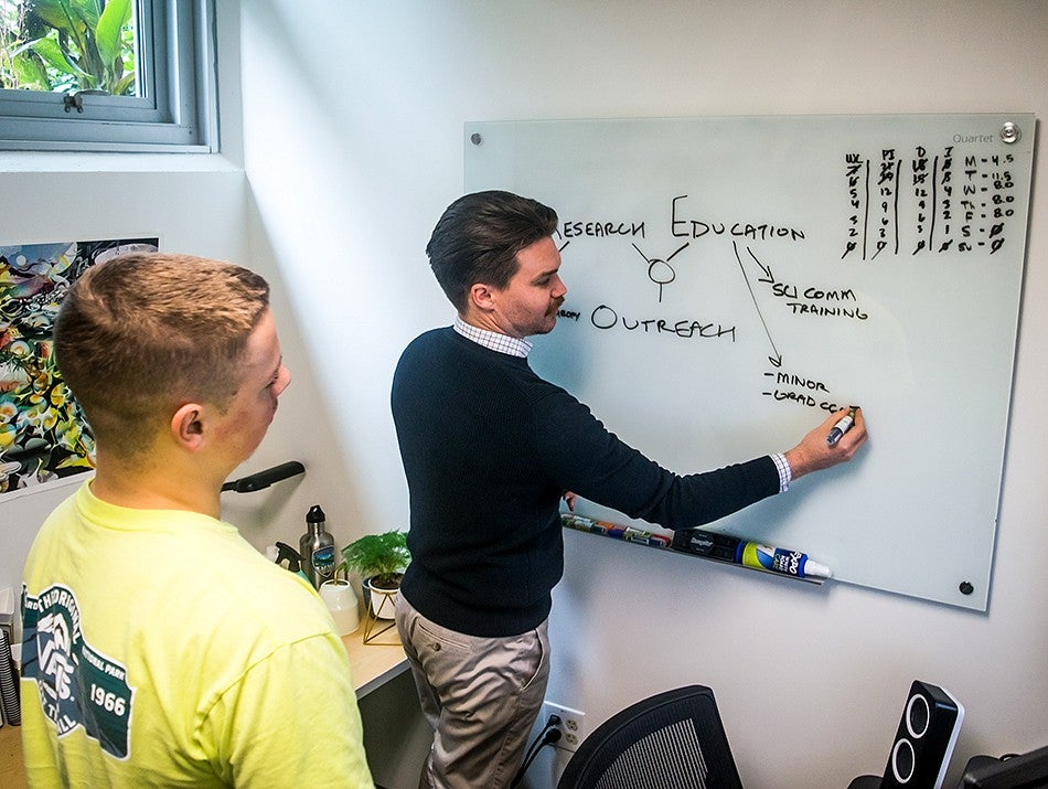 Two people work together in an office space. One person writes on a white board while the other person observes.