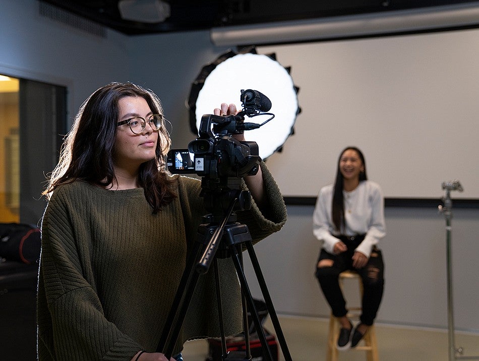 Two students work in the production studio. One student is seated in a stool, while the other student adjusts the settings on video camera.