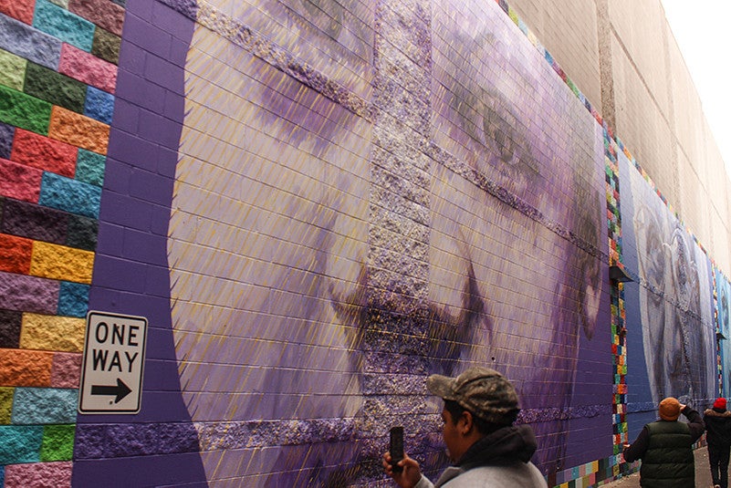 A person wearing a camouflage hat takes a photo of a purple mural. A person wearing a camouflage hat takes a photo of a purple mural.