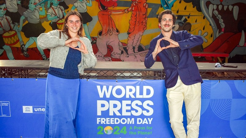Ruby Wool and Zachary Jones Neuray, student journalists representing the SOJC at the World Press Freedom Day conference, make O shapes with their hands while standing in front of a bright blue banner and a colorful mural