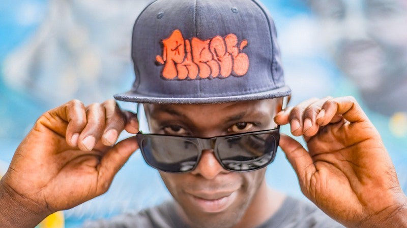 a person wearing a black hat with an orange graffiti-style imprint lowers his sunglasses down the bridge of his nose