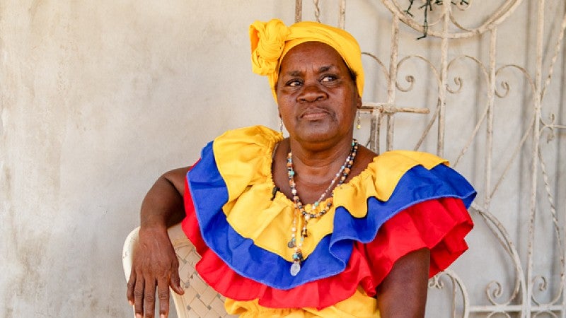 A woman wearing a vibrant dress inspired by the Colombian flag sits on a chair