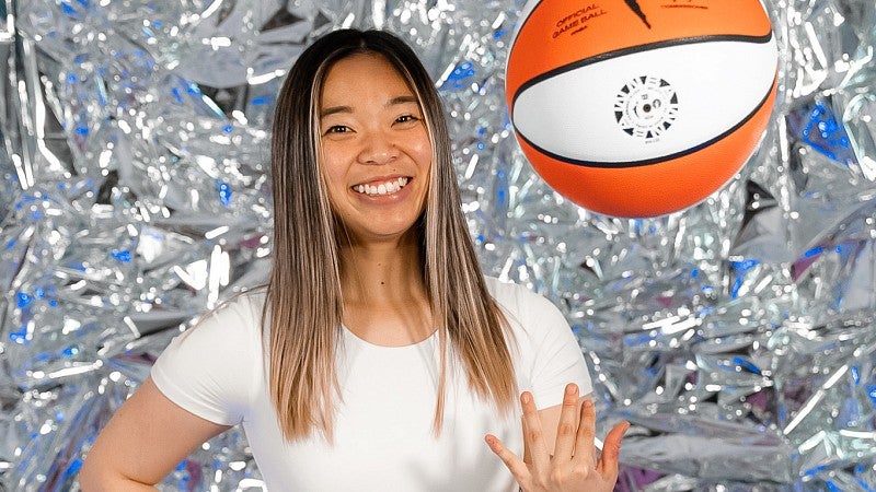 Carly Ebisuya poses for a portrait in front of a shiny silver background while tossing a basketball in the air 