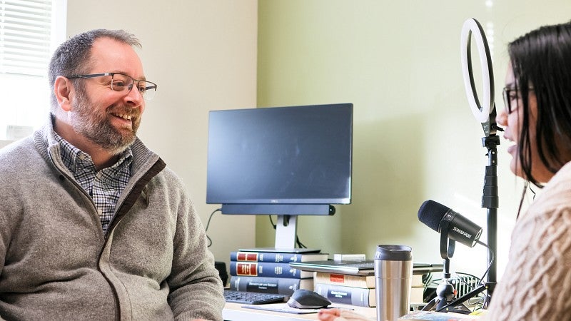 Bryce Newell sits at his desk and talks to a student