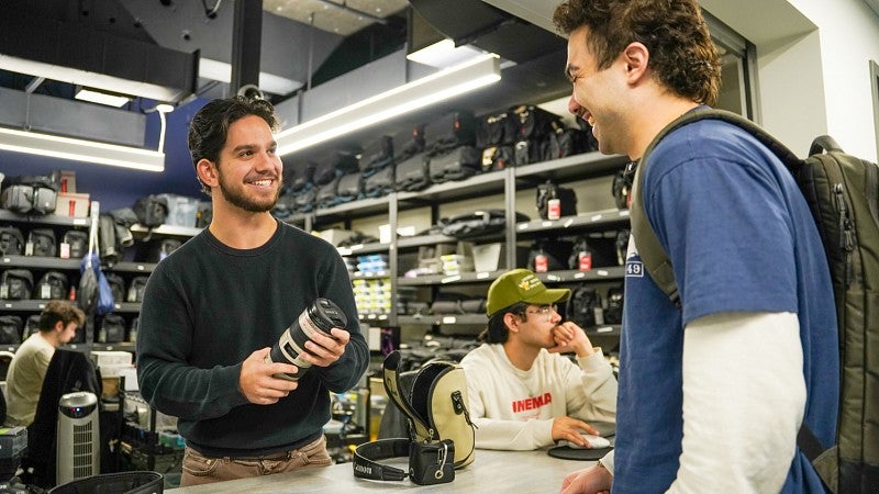 A student hands Zakary Christen-Cooney a camera lens from behind the desk of the SOJC J-Cage