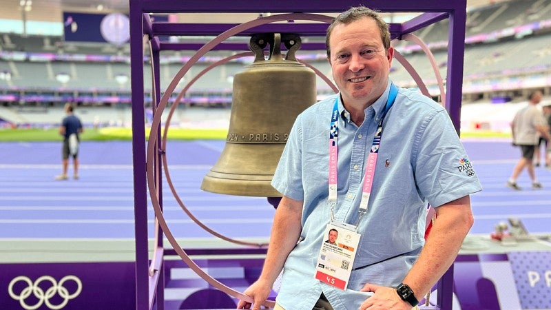 Paul Swangard poses with a large bell at the track and field stadium at the 2024 Paris Olympics