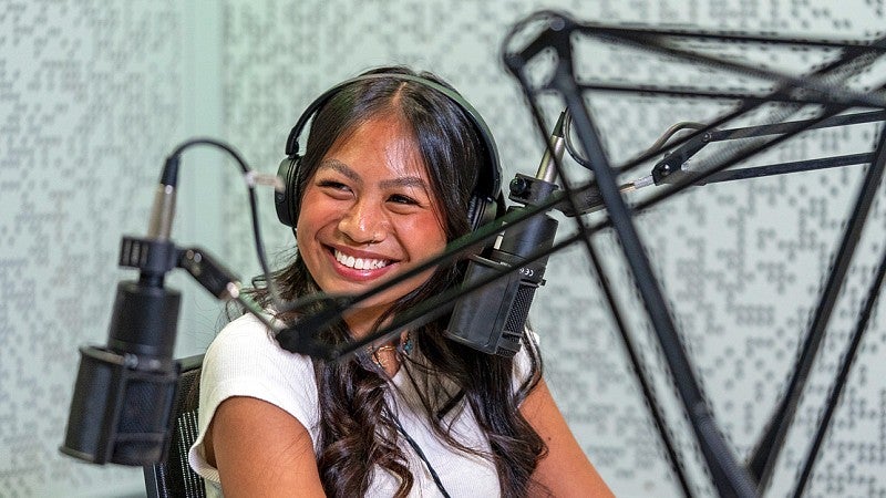 a student looks off camera and smiles while sitting in a podcast recording studio and wearing headphones