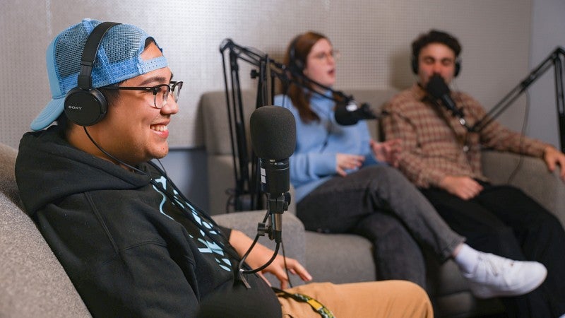 three students sit on couches in the University of Oregon School of Journalism and Communication's podcasting studio with microphones in front of them