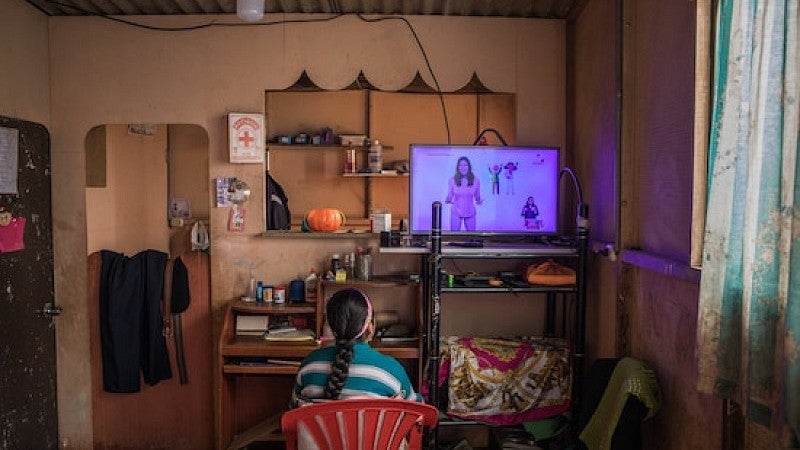 A peruvian student sits in a red plastic chair watching an educational broadcast at home while unable to attend school due to the COVID-19 pandemic. 