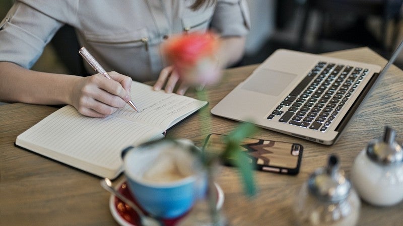 Person wearing a beige button down shirt writing in their notebook while sitting in front of a laptop.