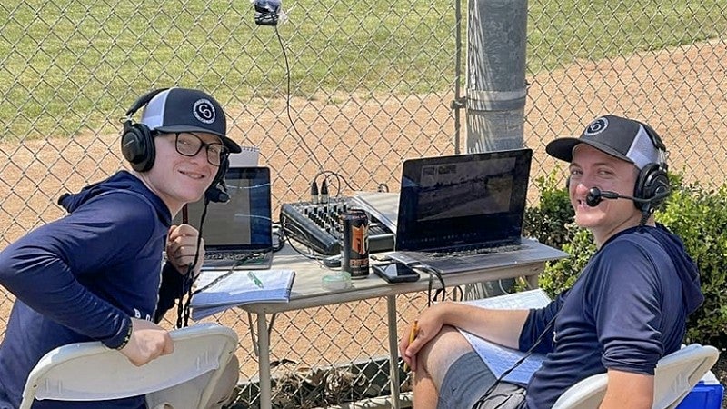 Two young men sitting at a table with sound equipment on the edge of a sports facility