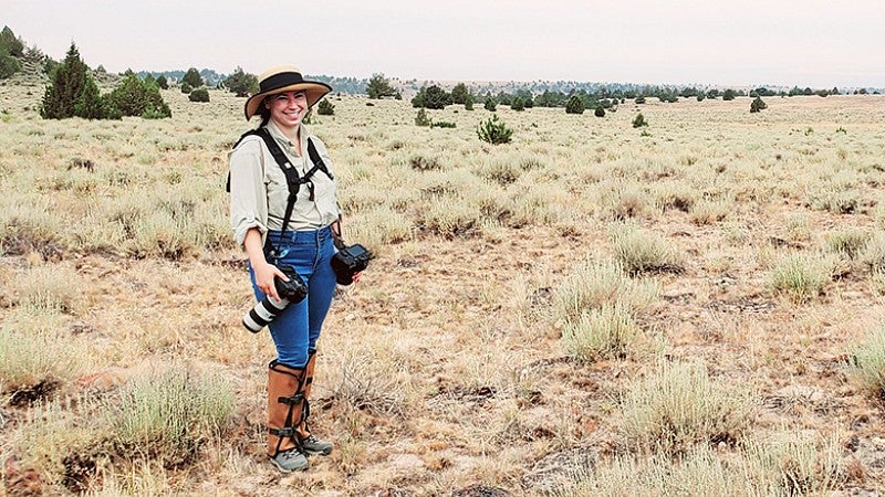 A young woman in hiking attire and holding camera equipment stands in an outdoor setting