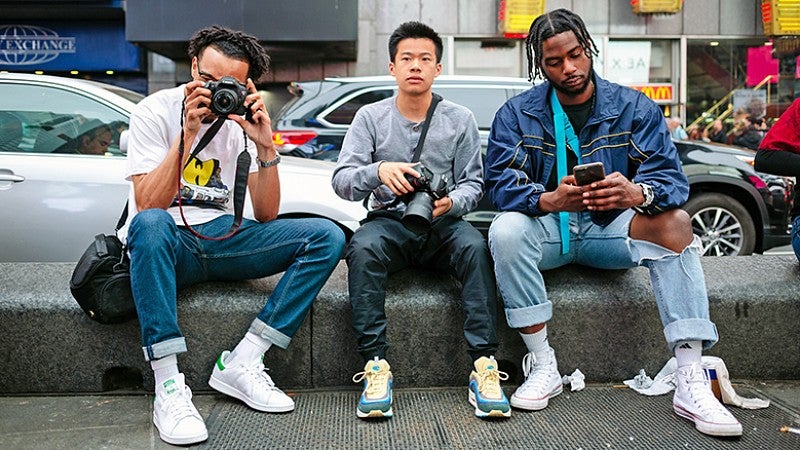 Three students in NYC sitting on curb holding cameras. 