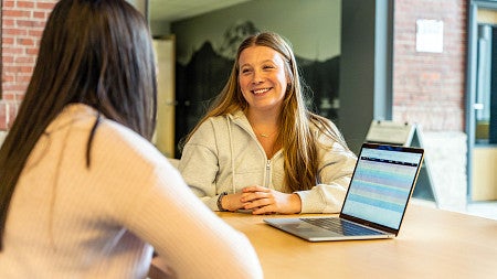 Erin Tooley chats with another student while they sit at a table with an open laptop in front of them