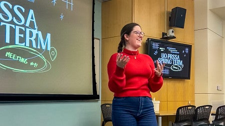 Jillian Gray wears a red sweater and gestures while presenting in front of a screen that says UO PRSSA Spring Term Meeting #1