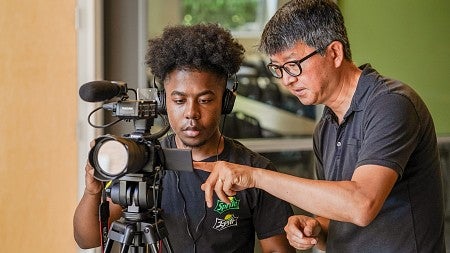 a student wearing headphones operates a video camera, while Sung Park points to the viewfinder screen