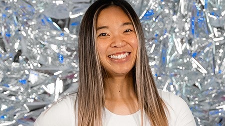 Carly Ebisuya poses for a portrait in front of a shiny silver background while tossing a basketball in the air 