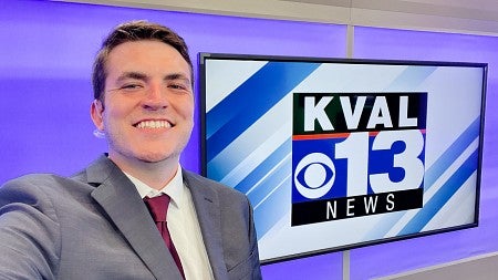 Billy Spotz takes a selfie in front of a screen displaying the KVAL 13 News logo