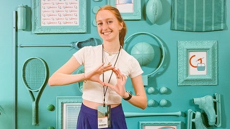 Maggie Troxell poses with her hands in the shape of an O in front of a wall of sports equipment painted teal at the Gondola sports content marketing conference