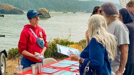 Sof Fox, a UO science communication minor, stands at a table laid out with informational materials with a river and pine forest in the background
