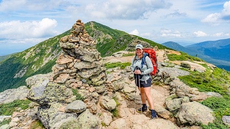 Denise Silfee wears backpacking gear and stands next to a large cairn with the peaks of mountains behind her