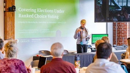 Regina Lawrence presents in front of a digital screen that says "Covering Elections Under Ranked Choice Voting"