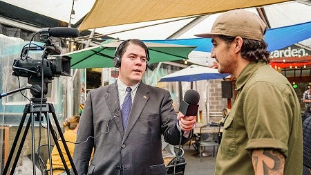 a student wearing a suit and holding a microphone interviews a person on camera