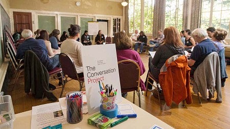 people gather in a circle of chairs with a sign in front that says "The Conversation Project"