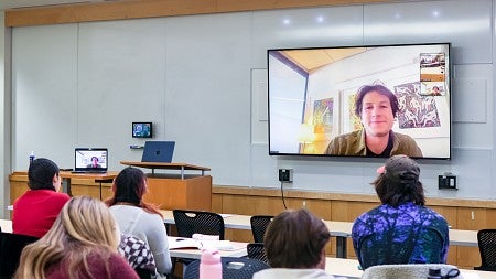 students face a display screen showing a video call