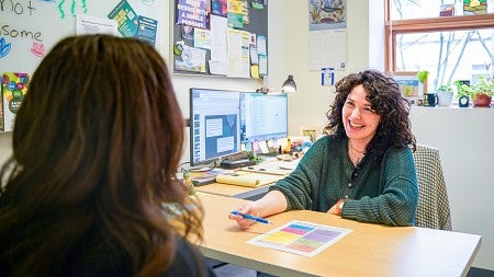 Annie Reiva smiles while sitting at her desk and talking to a student