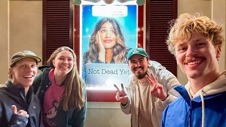 a group of people including Nolan Biorn pose in front of a poster for the show Not Dead Yet featuring the actress Gina Rodriguez