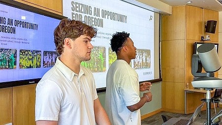 two students present in front of digital screens