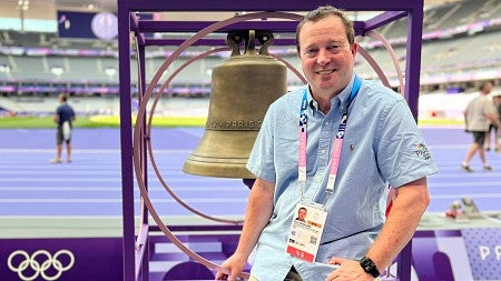 Paul Swangard poses with a large bell at the track and field stadium at the 2024 Paris Olympics