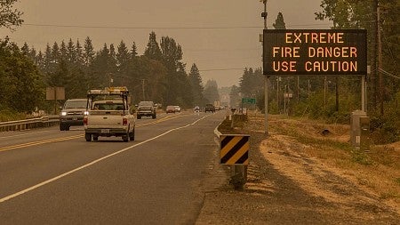 Cars drive along a highway during extremely smoky conditions; a sign next to the road reads "extreme fire danger, use caution"