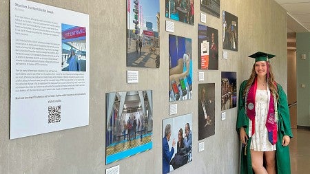 Ally Thomas, wearing her graduation cap and gown, stands next to her photo exhibit in Allen hall