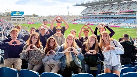 a group of University of Oregon School of Journalism and Communication students throw their Os as they pose in the stands of a large athletic stadium, with the field behind them