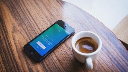 Cup of coffee on a wooden table next to a cellphone showing Twitter's login page.