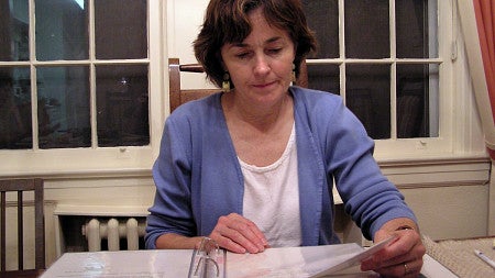 Faris reads Myra Genn's scrapbook while sitting at a table and wearing a lavender blouse. 