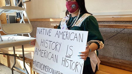 Angela Noah sits in the Many Nations Longhouse building holding a protest sign.